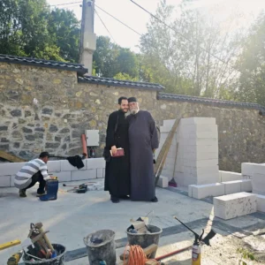 Doi clerici în rasă, în curtea Schitului Nașterea Maicii Domnului Chilii aflat în construcție, lângă zidul de piatră și blocurile de zidărie